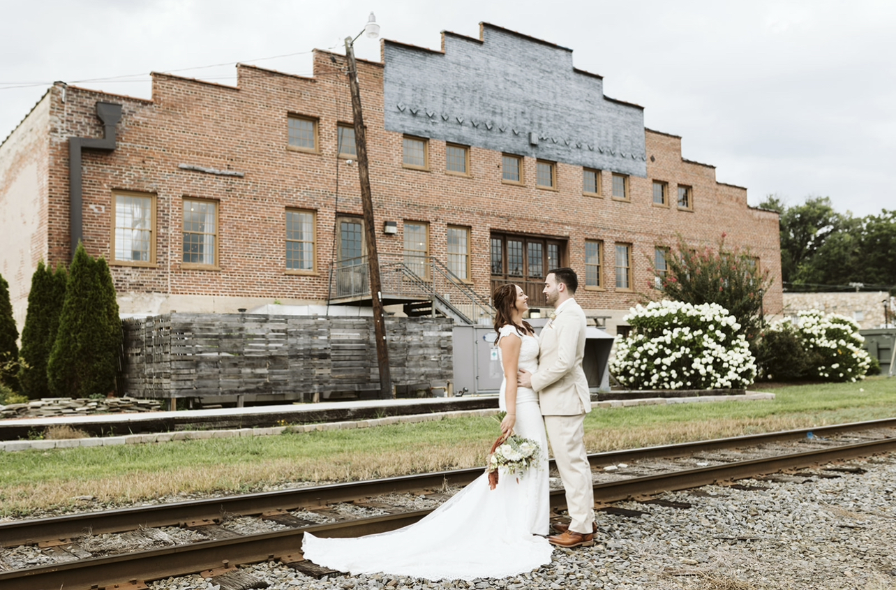 A couple shares a romantic moment on the railroad tracks behind Coley Hall at The Liberty, with a rustic brick building and vibrant landscaping in the background.