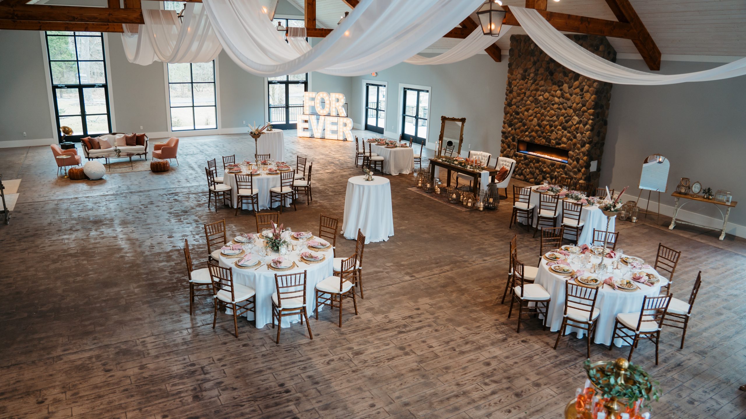 A beautifully decorated reception hall at White Sulphur Springs, featuring draped white fabric, round tables with elegant table settings, and a cozy stone fireplace. Photo by Dakota Goss Photography.