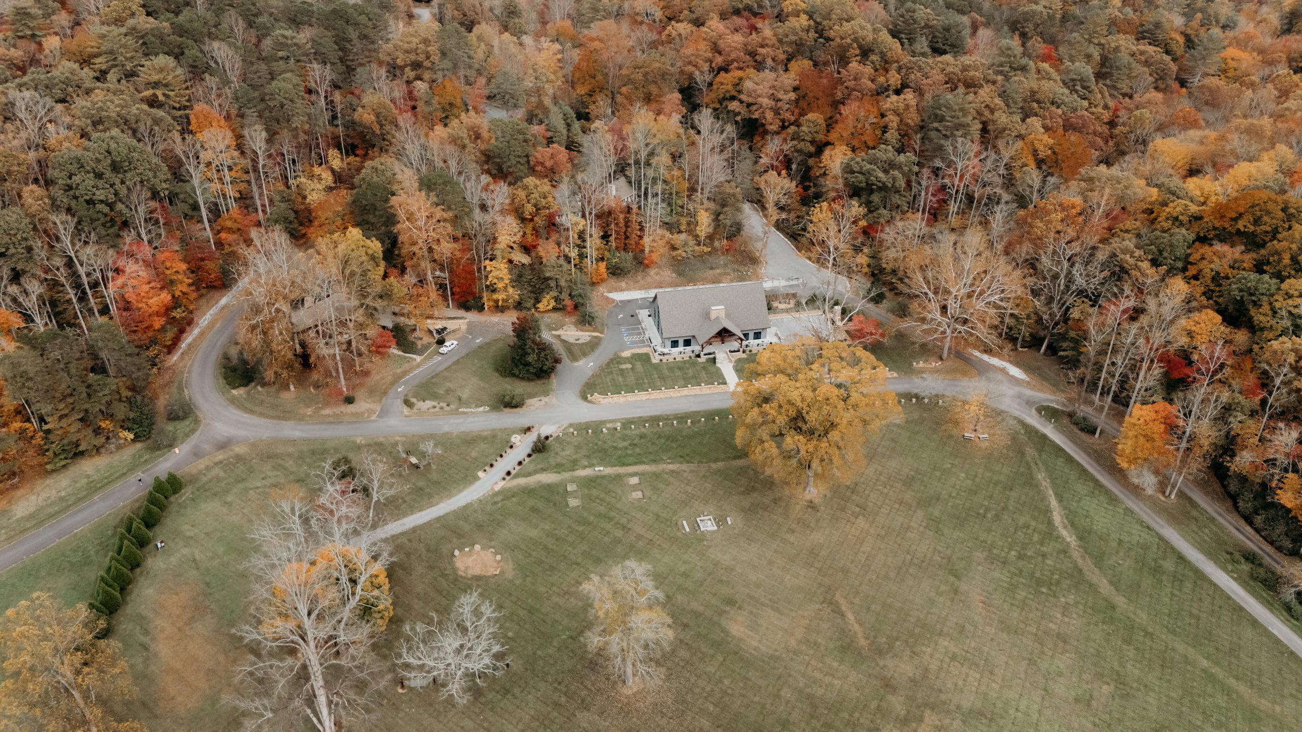 A stunning aerial view of the Lodge Venue at White Sulphur Springs surrounded by vibrant autumn foliage, showcasing the expansive grounds with winding paths, open fields, and the lodge nestled among the trees.