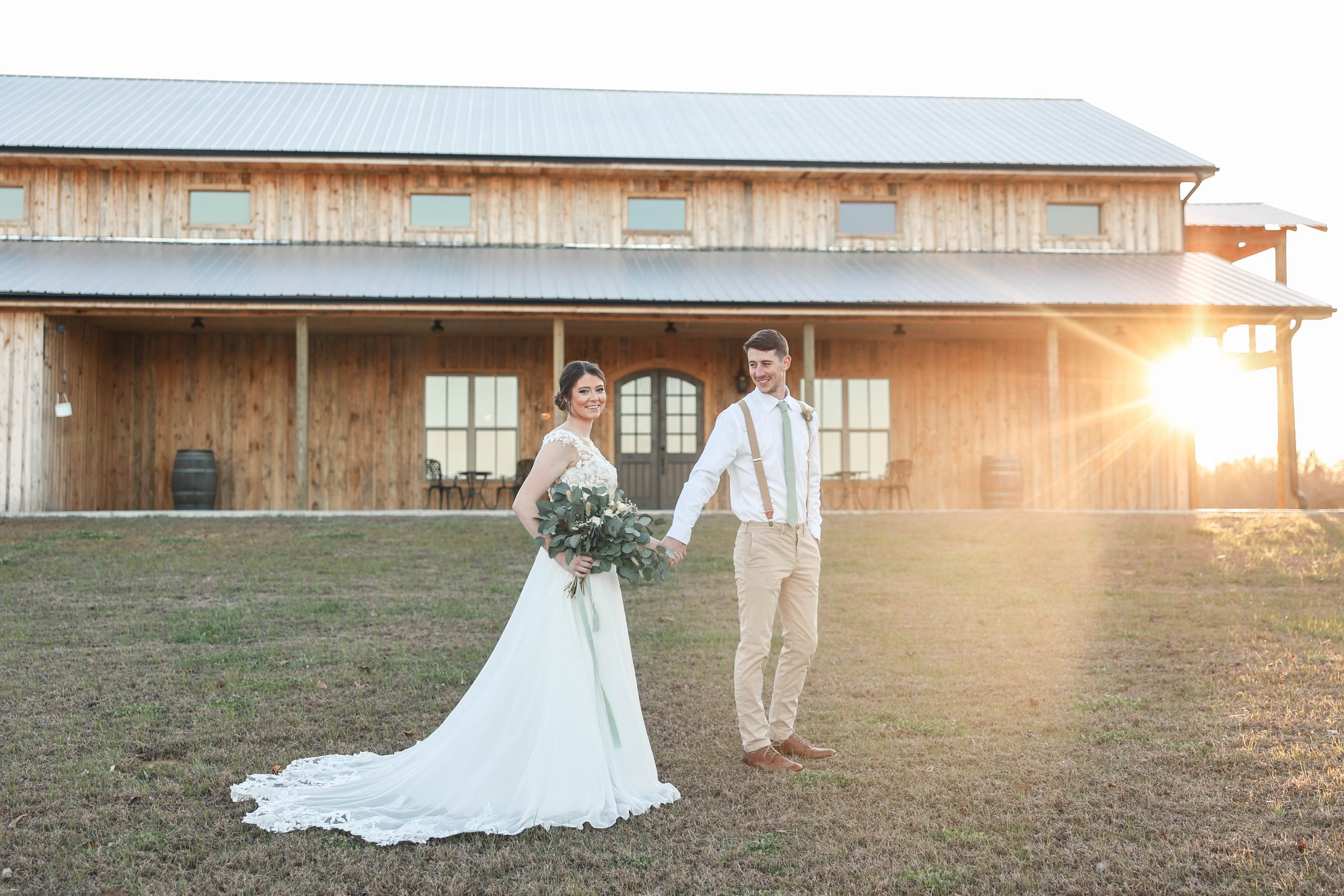 Bride and groom holding hands in front of The Barn at Premier Farm, a rustic wooden barn with warm sunlight streaming in the background. Captured by Little Wagon Photography.