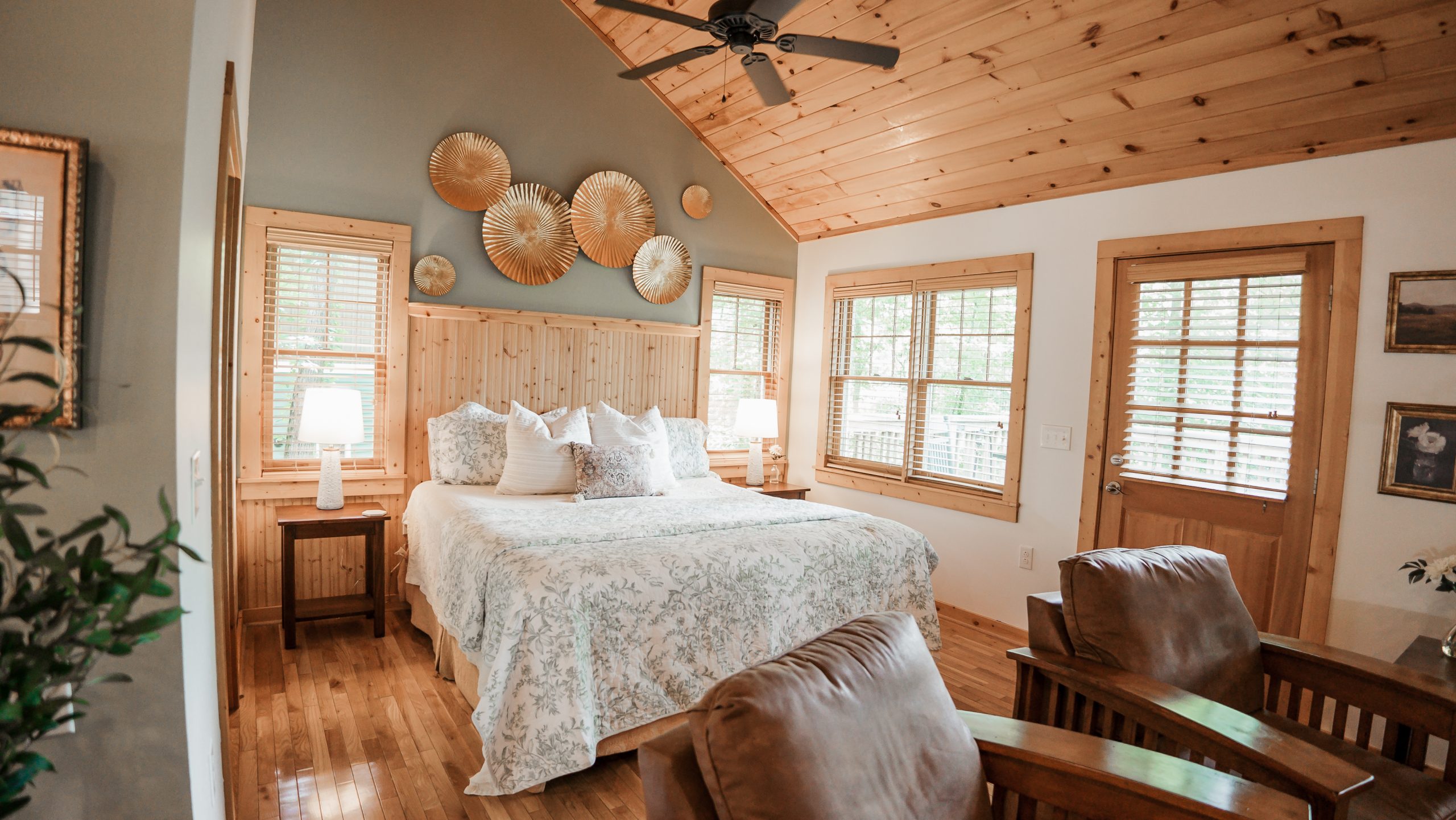 The interior of a cozy cabin at White Sulphur Springs, showcasing a charming wooden ceiling, a comfortable bed with floral bedding, and natural lighting from large windows. Photo by Dakota Goss Photography.