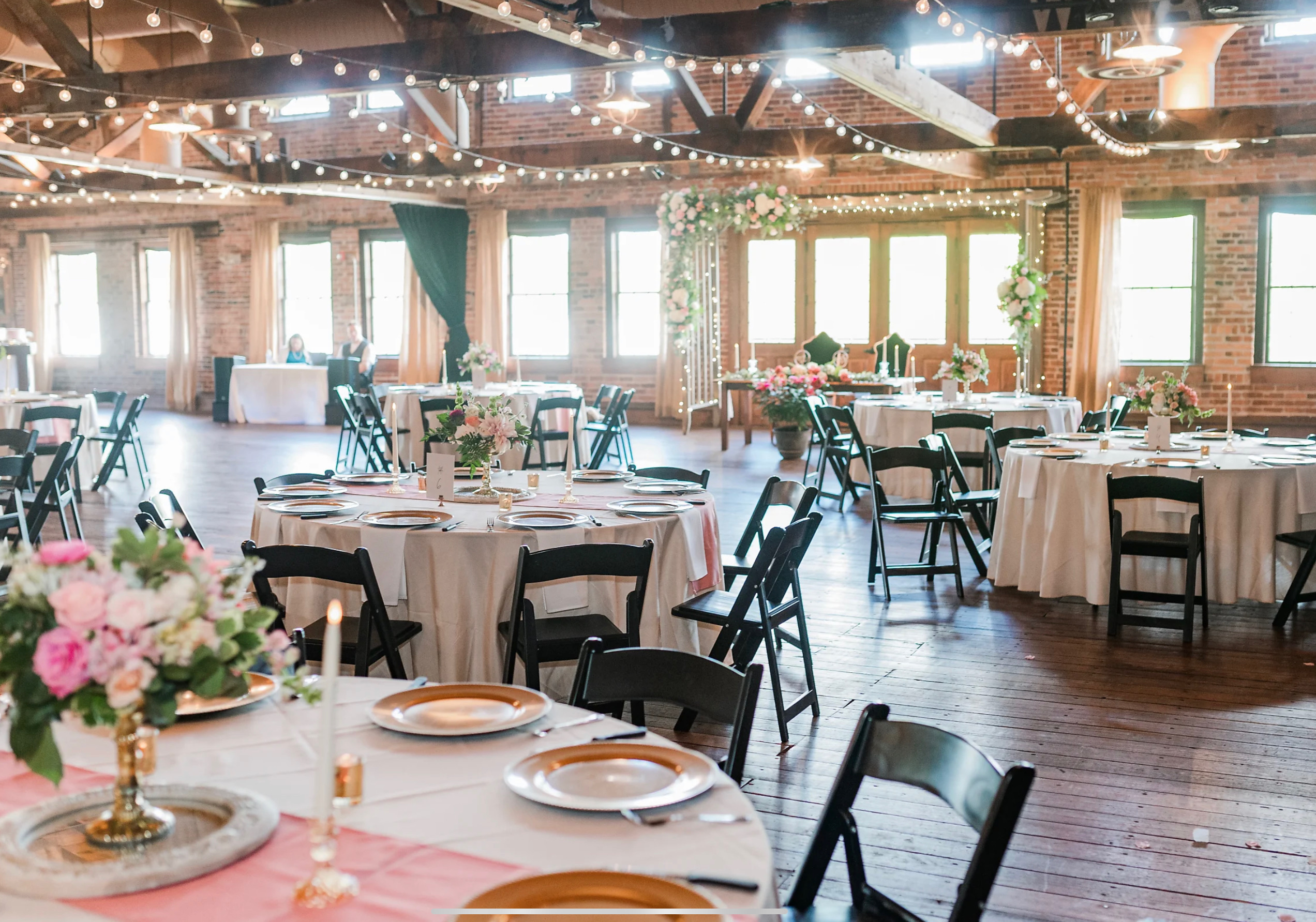  A beautifully arranged reception space at Coley Hall at The Liberty, featuring round tables set with pink and gold decor and surrounded by black chairs. The ambiance is enhanced by exposed wooden beams and warm string lights.