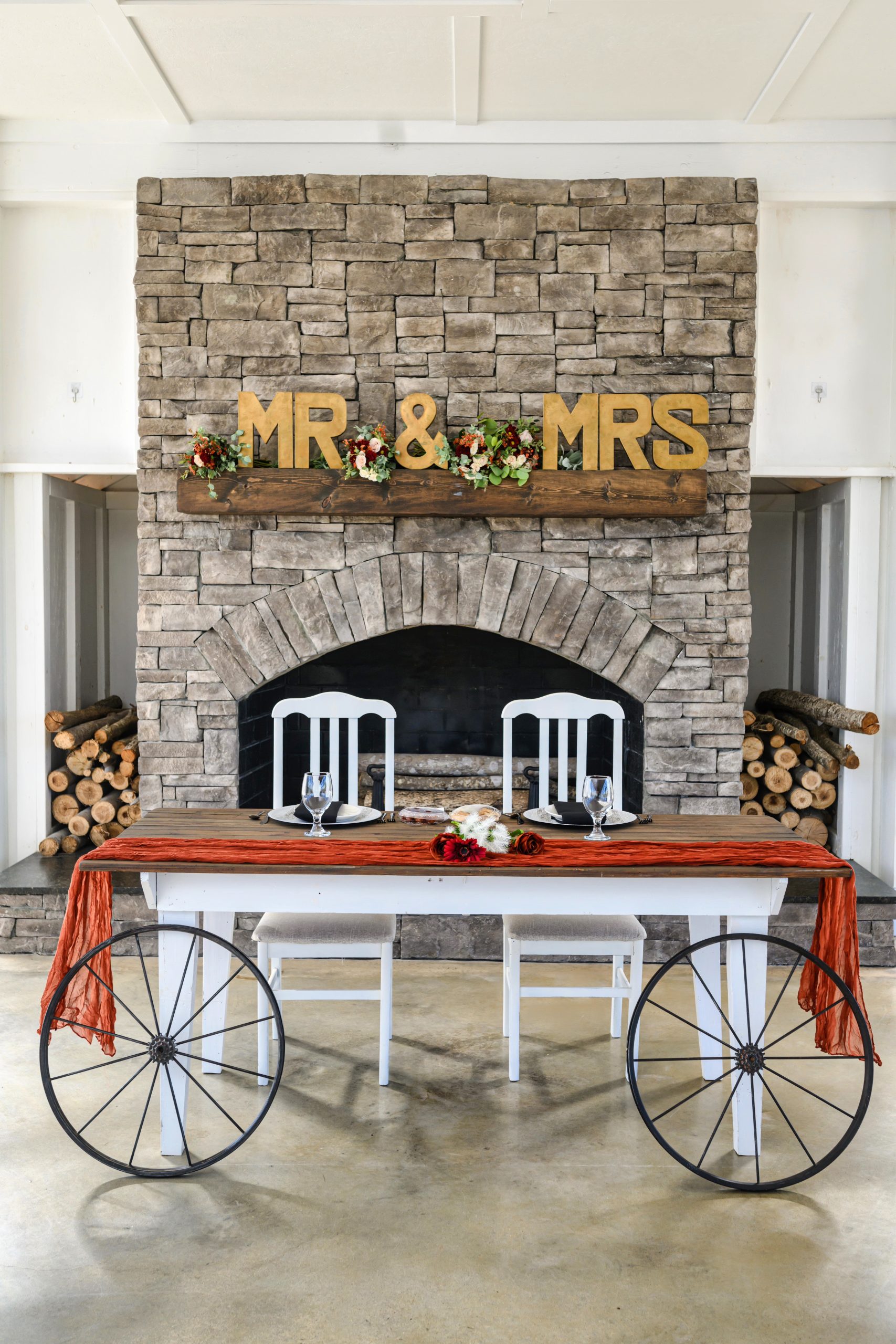 A rustic sweetheart table with wagon wheels and a wooden beam mantle decorated with "Mr & Mrs" letters and floral arrangements, placed in front of a stone fireplace.