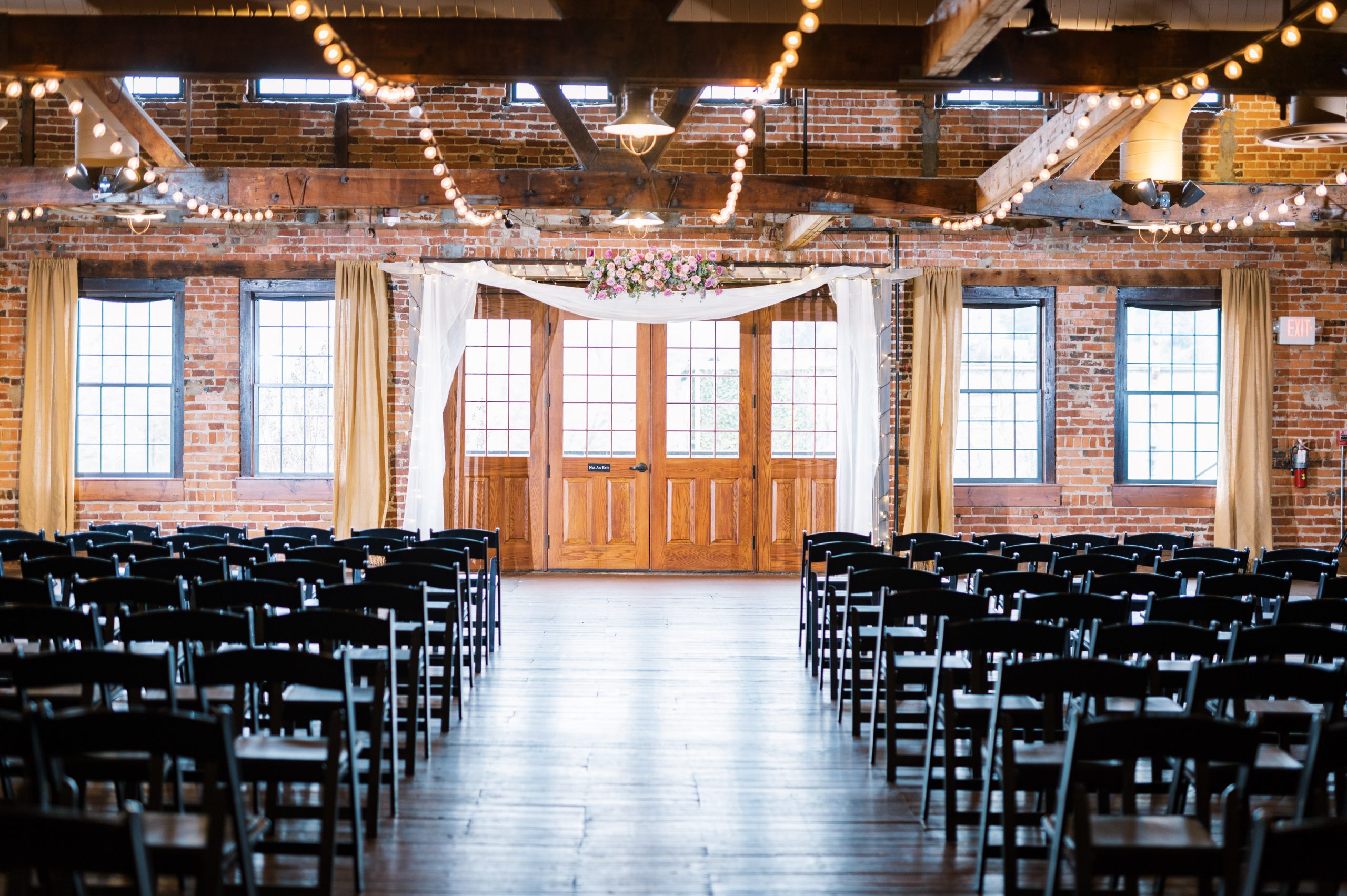 A romantic indoor ceremony setup at Coley Hall at The Liberty, with rows of black chairs facing a white floral arch and wooden double doors framed by sheer curtains. The space exudes a warm rustic charm with its exposed brick walls and glowing string lights.
