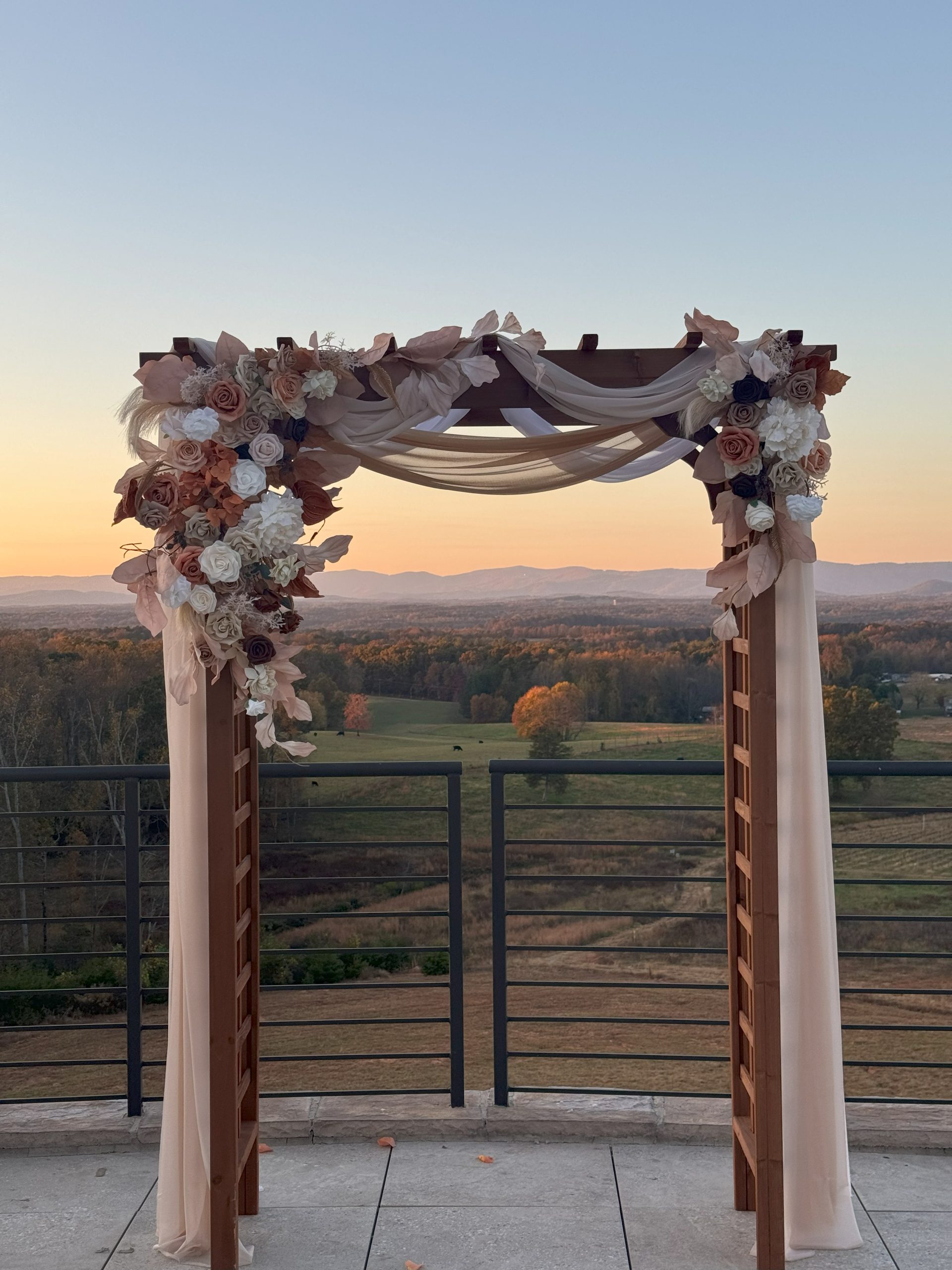 Wooden ceremony arch adorned with soft draping and floral arrangements in blush and cream tones, set against a panoramic sunset view of rolling hills at La Collina at Piccione Vineyards. Captured by Lauren McGlamery.