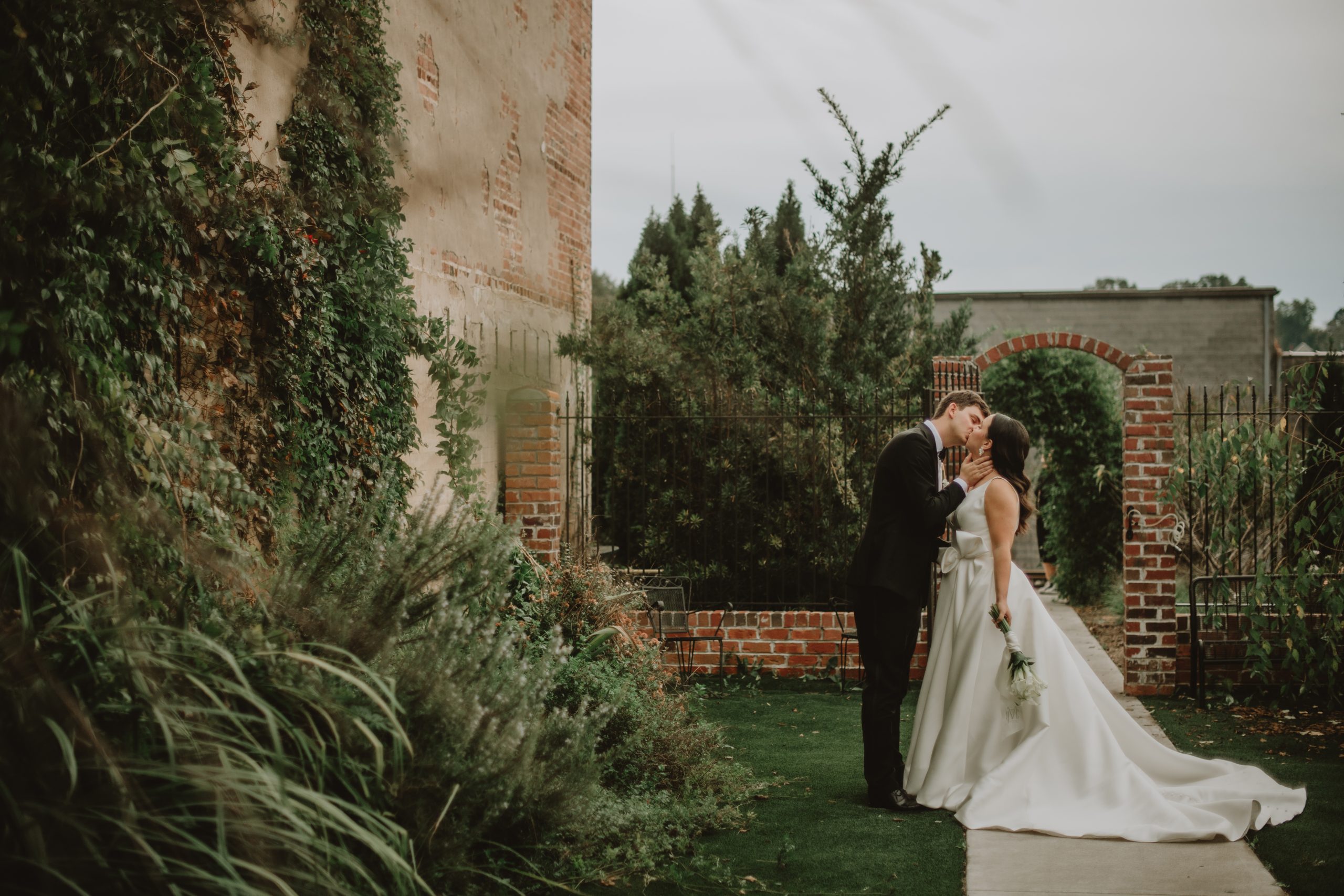 A bride and groom sharing a kiss in the Courtyard Garden at Coley Hall at The Liberty. The charming setting features lush greenery, brick walls, and an arched iron gate, creating a serene backdrop for the couple's intimate moment.