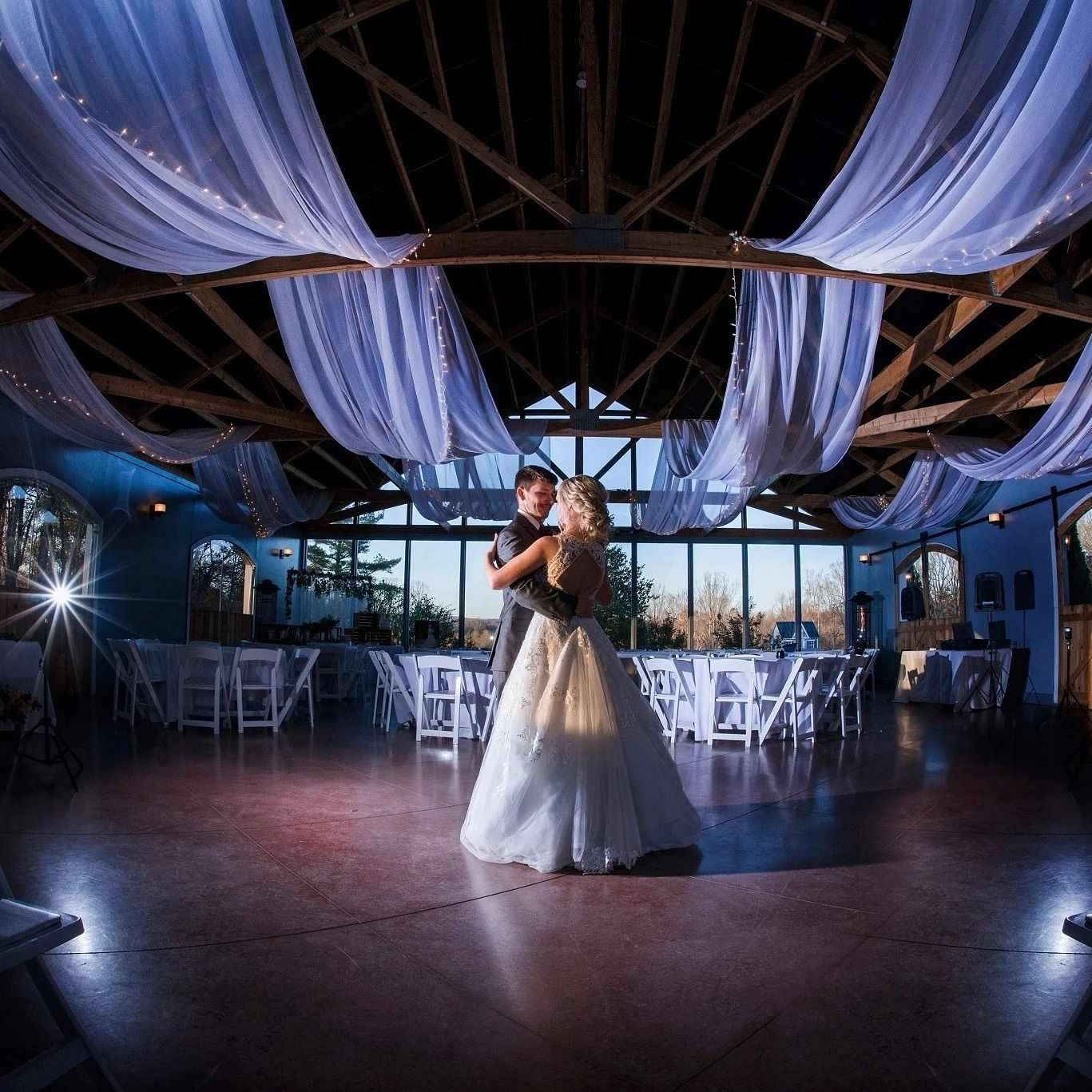 A romantic moment of a couple dancing in the Carriage House at Rosa Lee Manor, adorned with flowing white drapes, exposed wooden beams, and ambient natural light filtering through large windows.