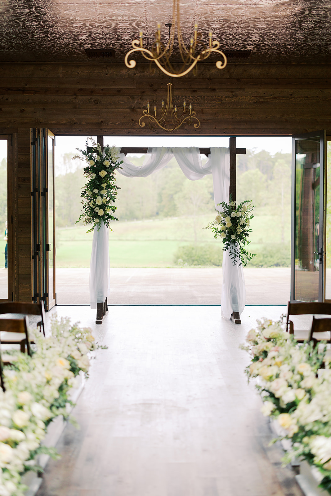 The elegant ceremony site at Luna's Trail Farm, featuring a wooden arch draped with white fabric and adorned with lush white floral arrangements, surrounded by a scenic outdoor backdrop. Photo by Alexandra Scott Photography.