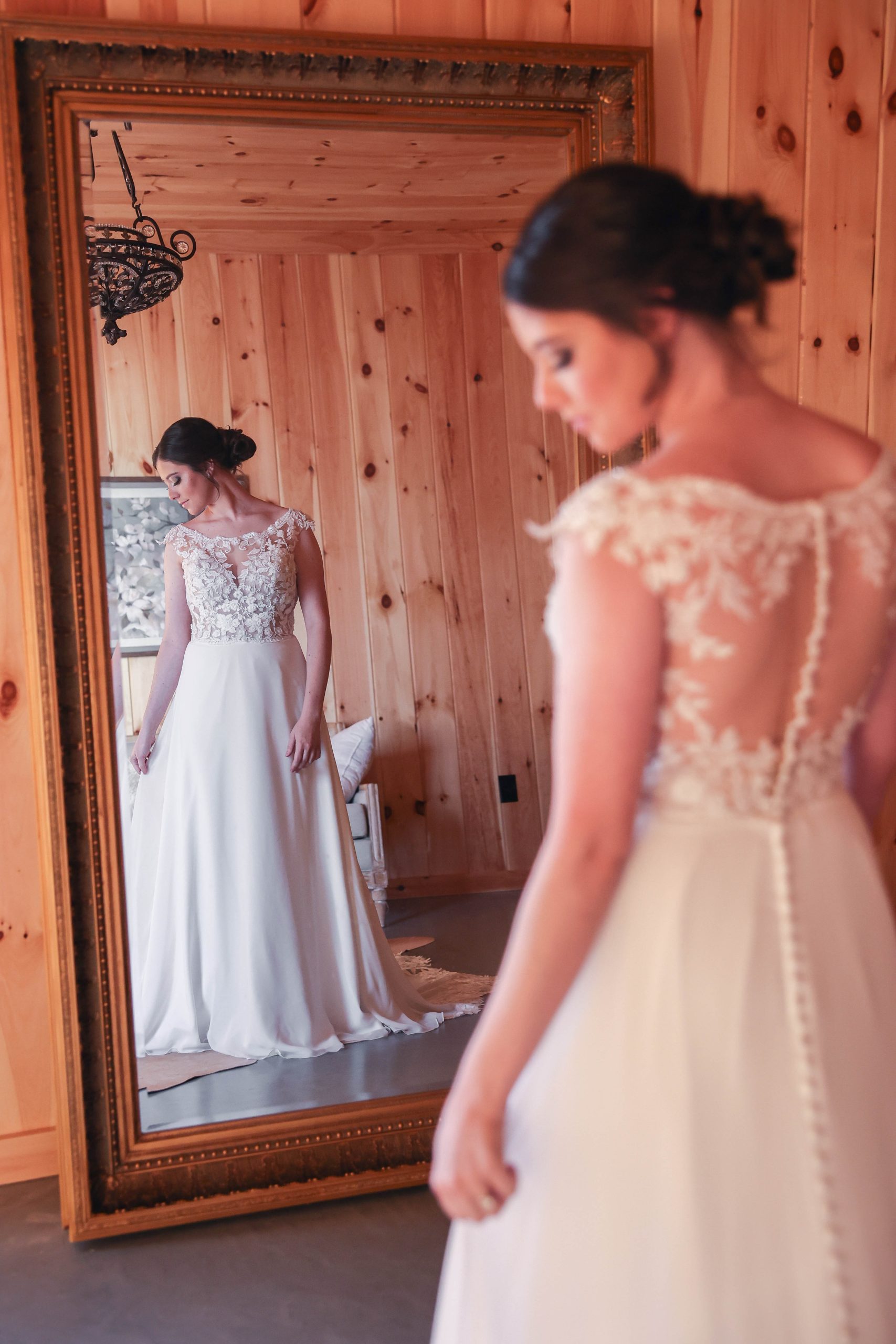 A bride standing in a cozy dressing suite with warm wooden walls, gazing at her reflection in an ornate full-length mirror while wearing an elegant lace wedding gown.