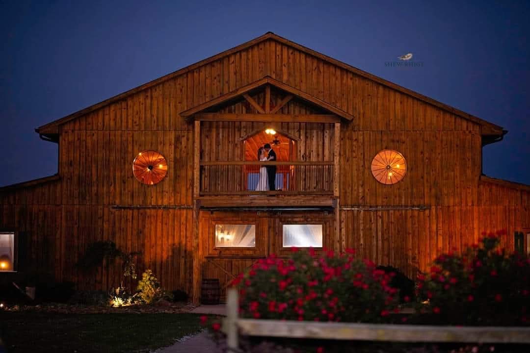 The Barn at Heritage Farm illuminated at night with warm lighting, featuring a couple standing on the balcony under a starlit sky. Captured by Shew Ridge Photography.