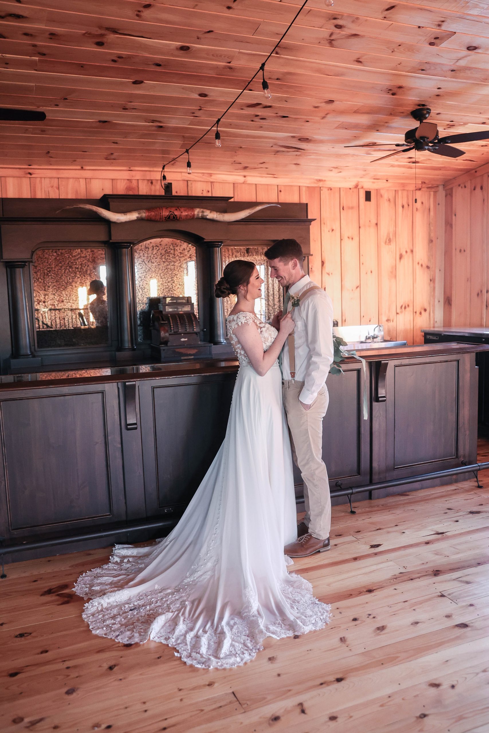 A bride and groom share a romantic moment in a lofted barn area with rustic wooden walls and a vintage-style bar, illuminated by warm string lights.