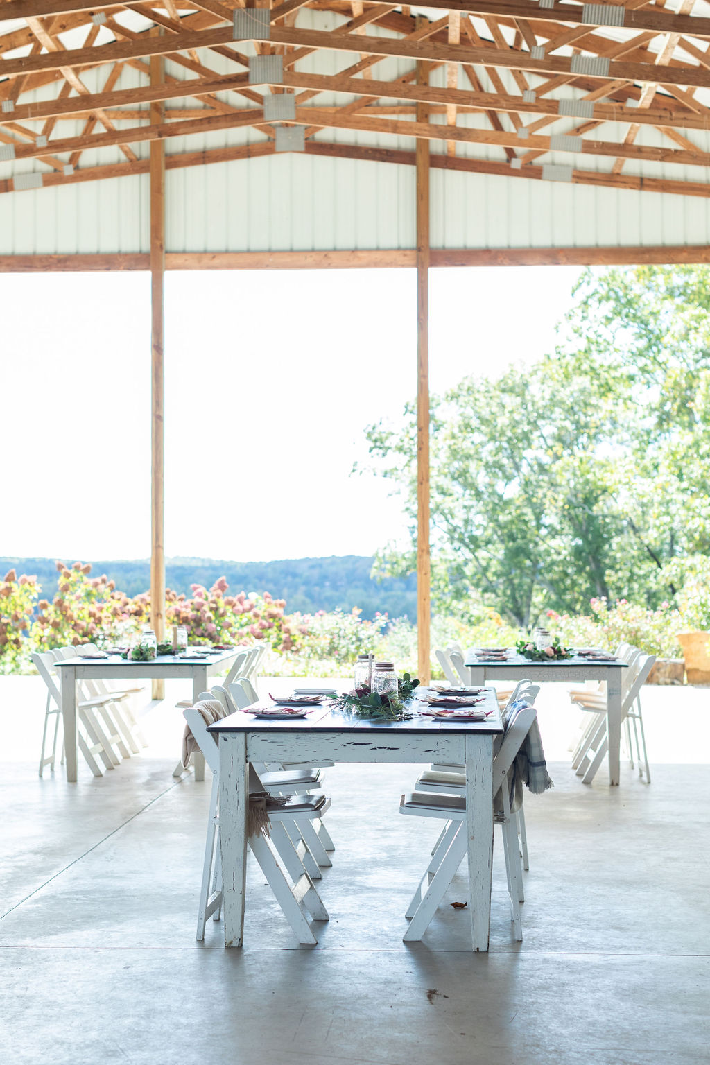 An open-air pavilion featuring rustic white tables set with greenery and floral centerpieces, overlooking a scenic view of rolling hills and blooming hydrangeas under a wooden beam ceiling.