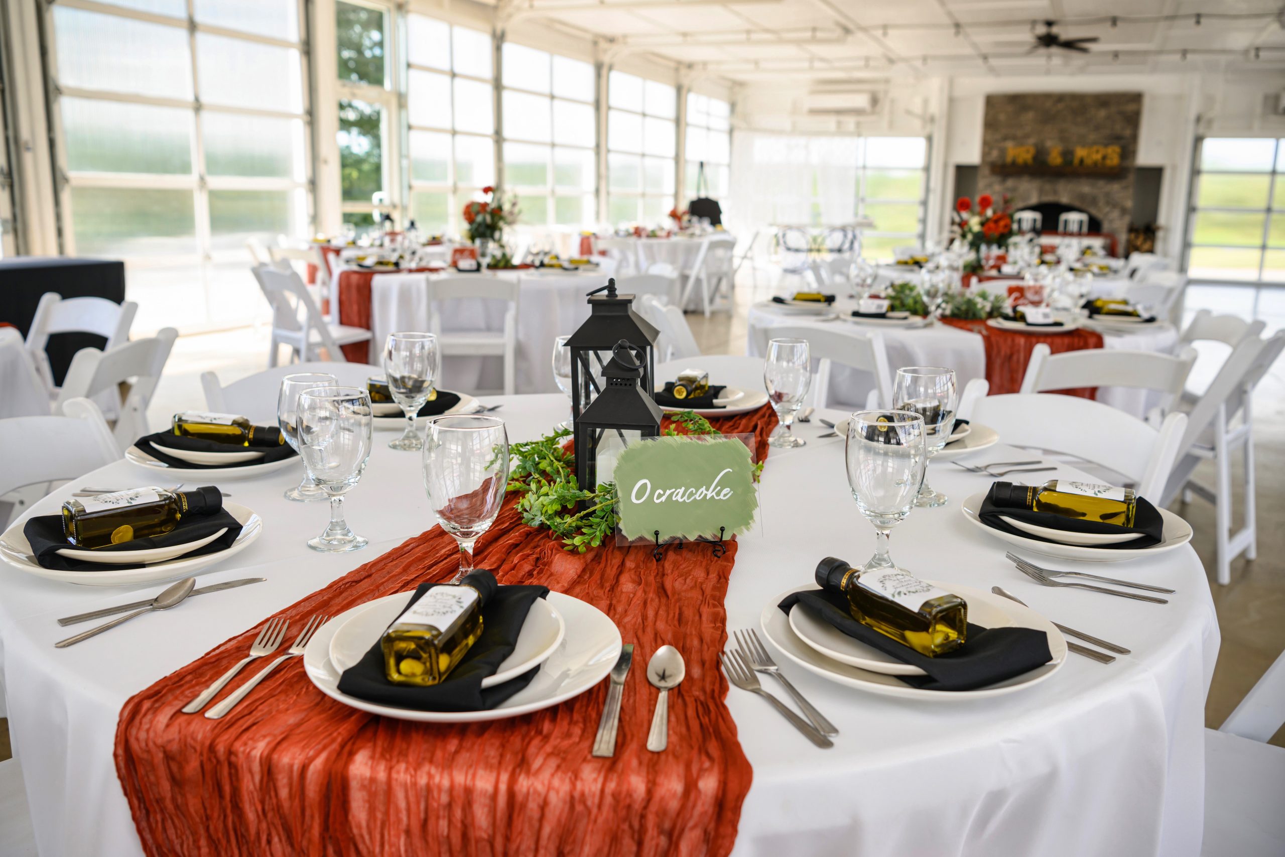 A beautifully arranged reception table at the pavilion in Mayberry Meadows, featuring a rustic orange table runner, mini olive oil bottle favors, elegant glassware, and a lantern centerpiece with greenery, set against a backdrop of bright, natural light.