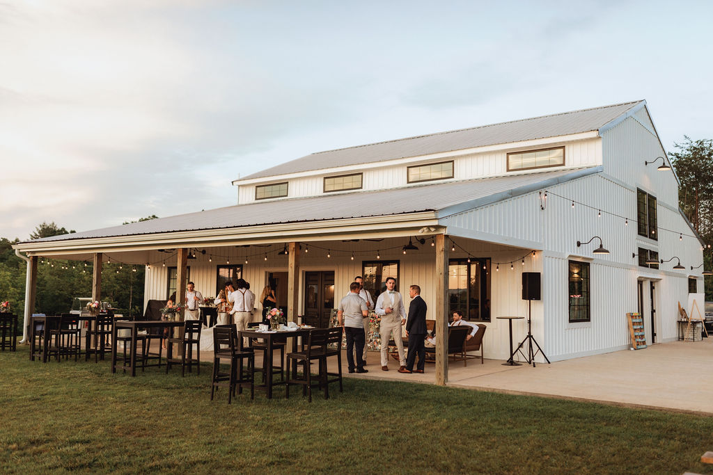 The porch at Serre Vineyards featuring outdoor seating, string lights, and a group of guests mingling at sunset. The modern white barn provides a stunning backdrop. Captured by Lee Bottoms Photography.