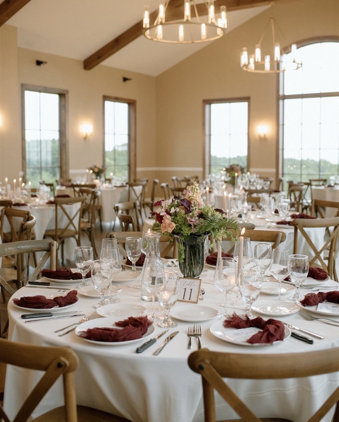Elegant reception hall at La Collina at Piccione Vineyards featuring round tables with white linens, burgundy napkins, floral centerpieces, and chandeliers under exposed wooden beams. Captured by Yaseli Olivera Photography.