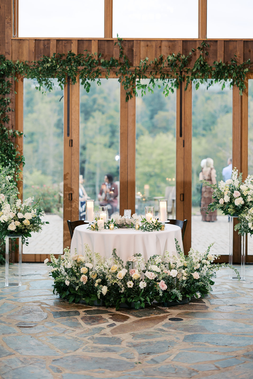 A beautifully decorated sweetheart table at Luna's Trail Farm reception hall, featuring cascading floral arrangements and greenery in front of tall windows overlooking the scenic outdoors.