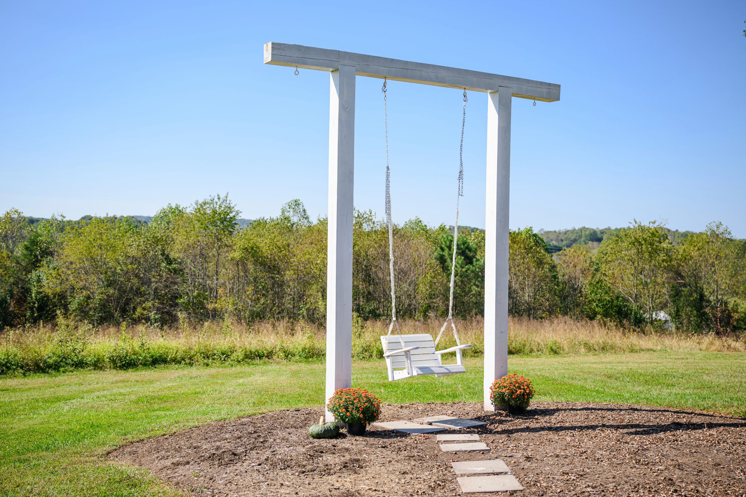 A white wooden swing adorned with a natural backdrop of greenery, placed on a circular patch of mulch surrounded by vibrant fall flowers.