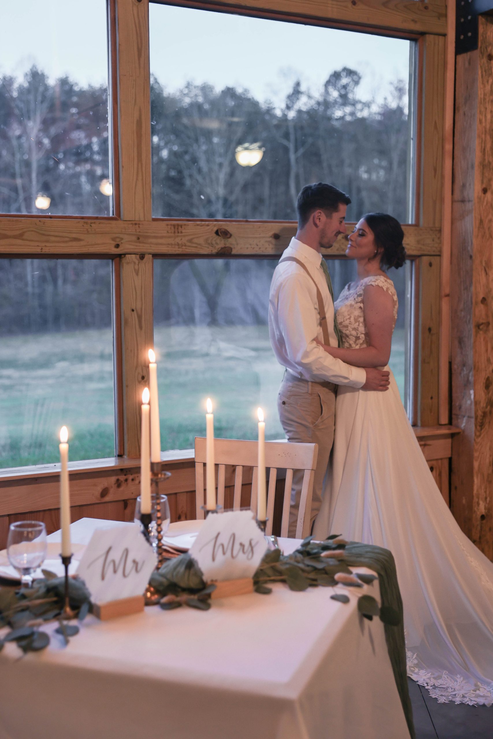 A bride and groom standing close by a sweetheart table adorned with candles and greenery, backlit by the soft glow of large floor-to-ceiling windows showcasing a serene outdoor view.