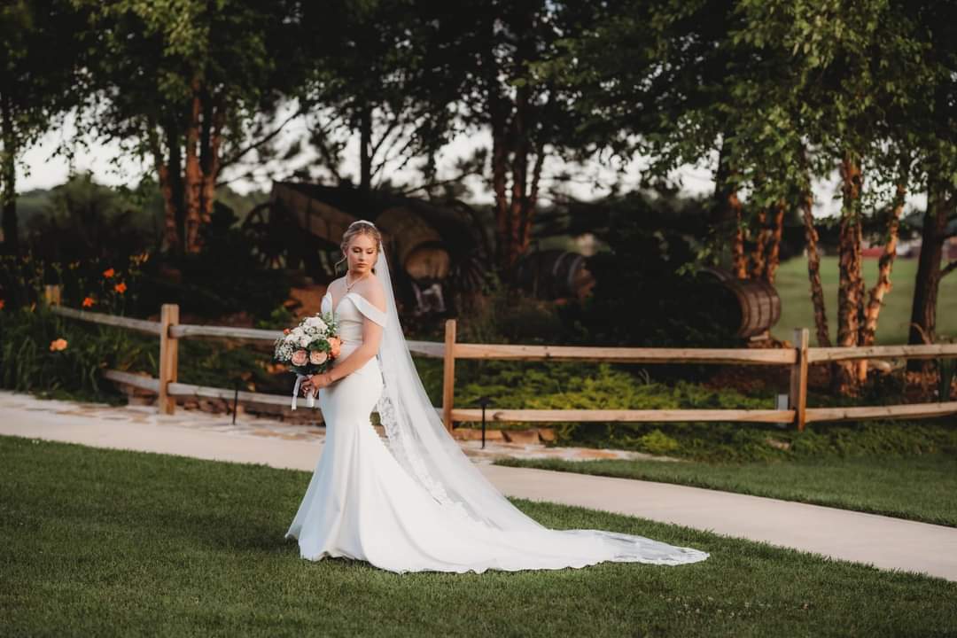 Bride standing gracefully on the lawn at The Barn at Heritage Farm, holding a bouquet of pink and white flowers with a rustic wooden fence and lush greenery in the background. Captured by Shew Ridge Photography.