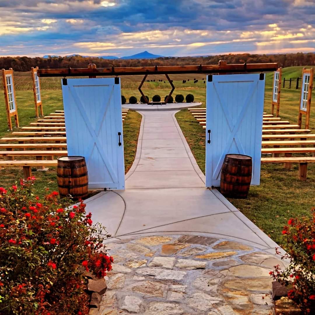 The Outdoor Chapel at The Barn at Heritage Farm, featuring a stone pathway, white barn doors, and a hexagonal arbor with a backdrop of rolling hills and vibrant skies at sunset.