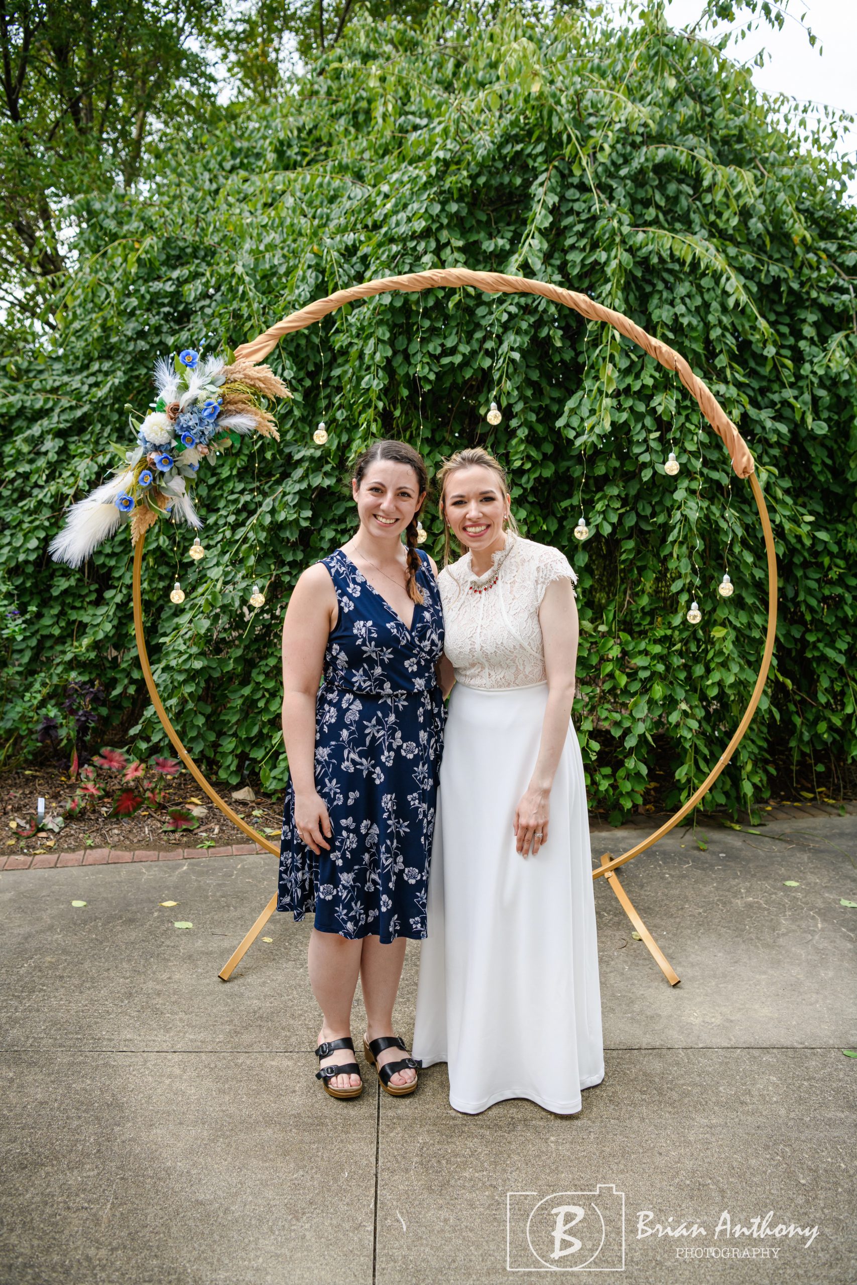 Bride Paula smiling alongside her planner Stephanie under a whimsical circular floral arch during her eclectic tea party wedding at JC Raulston Arboretum.