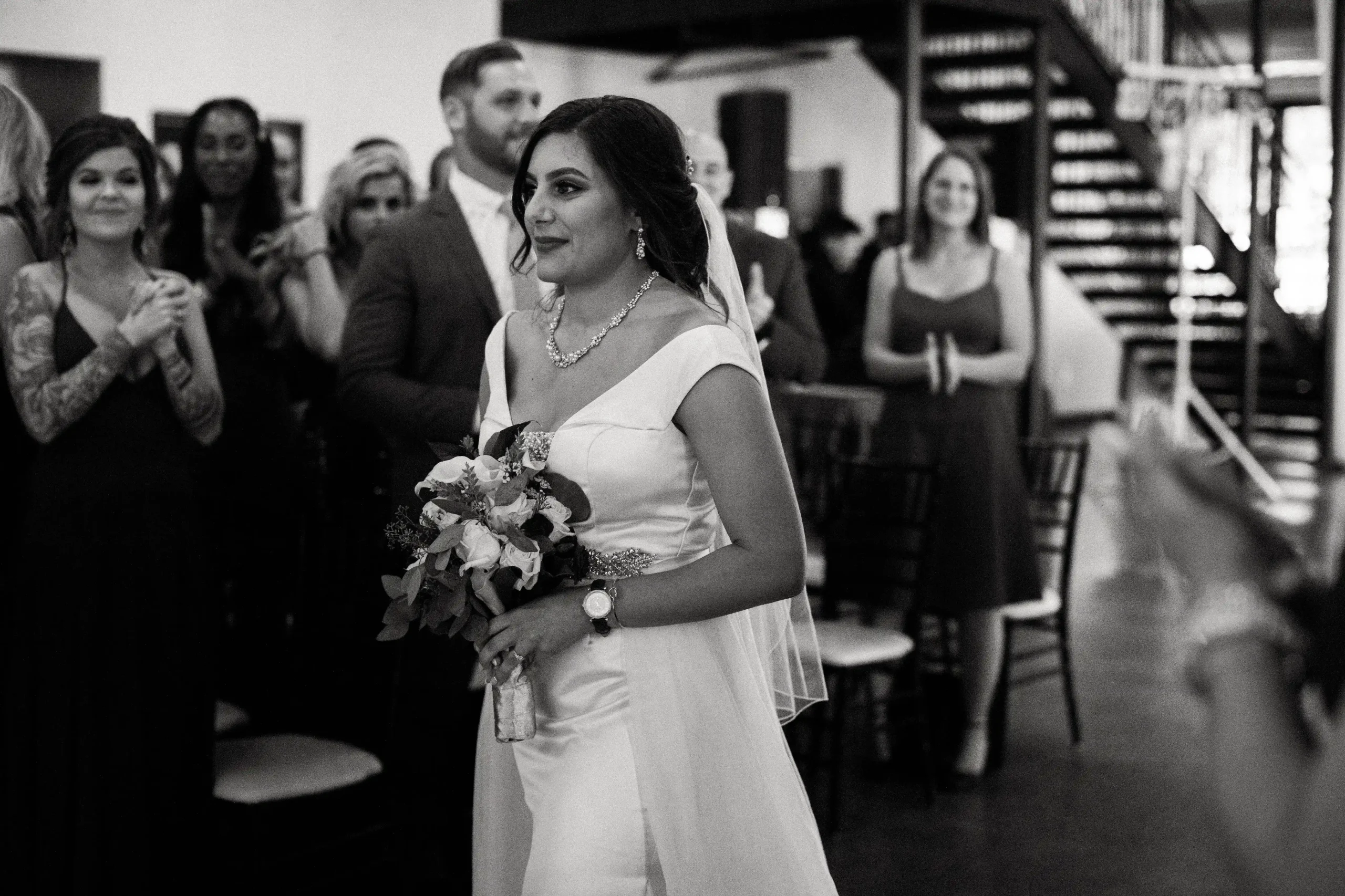 Black and white photo of a bride in a fitted gown holding a bouquet as she walks down the aisle alone, surrounded by applauding guests.