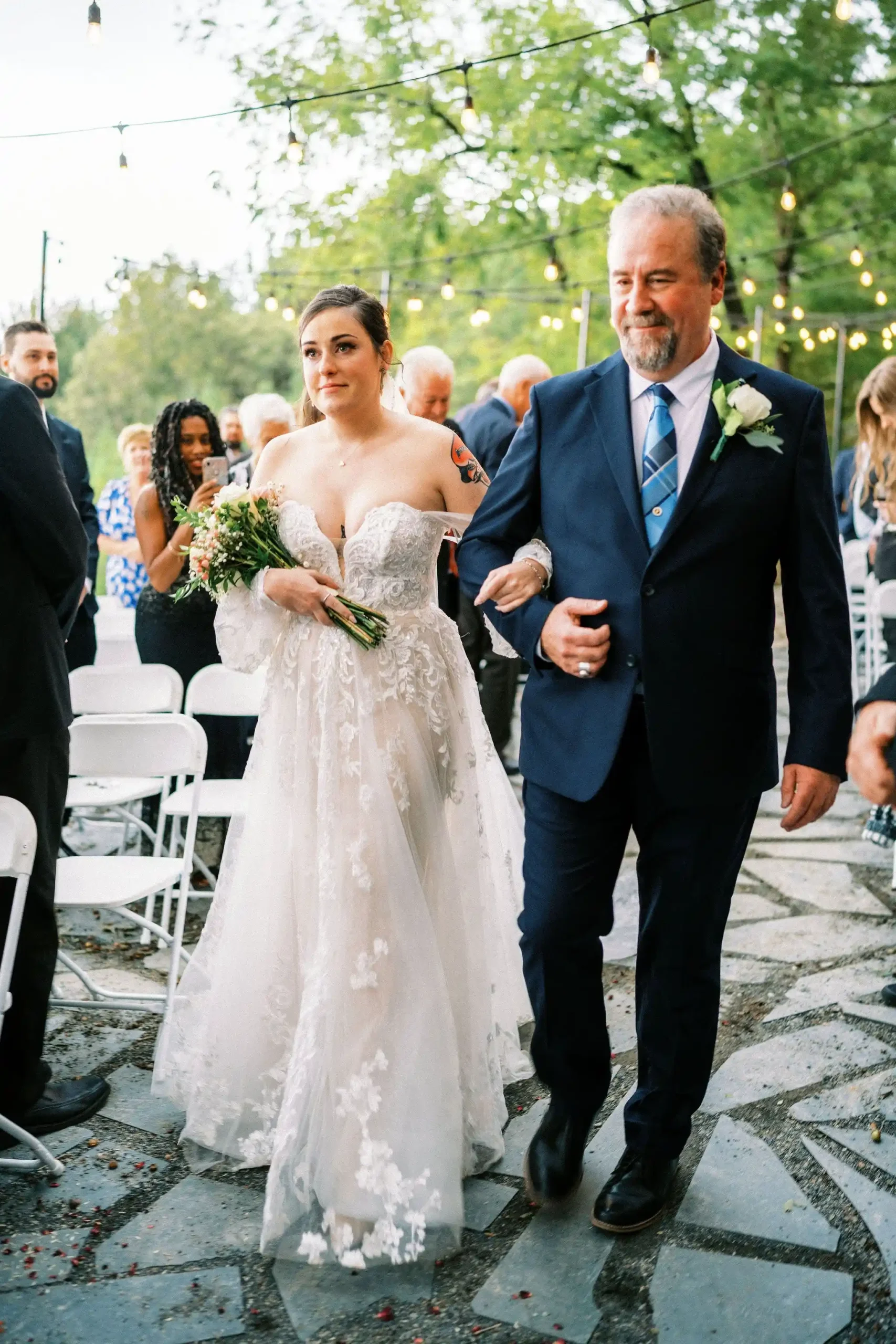 Bride in a lace wedding gown walks down the aisle with her father during an outdoor ceremony decorated with string lights.