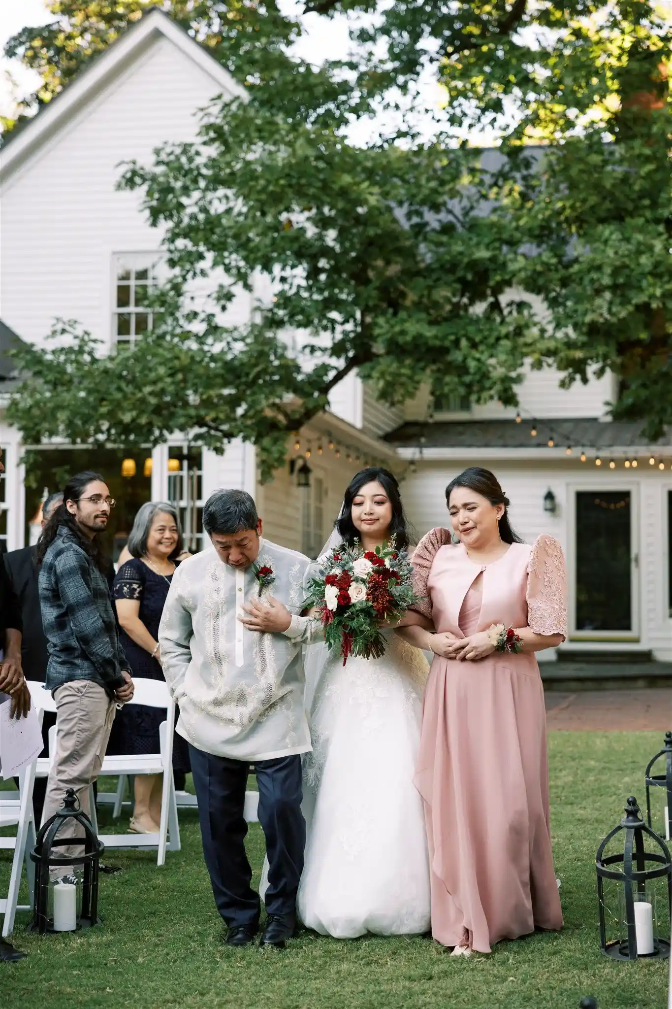Bride in a white gown holding a red and white bouquet walks down the aisle with her parents during an outdoor wedding ceremony.