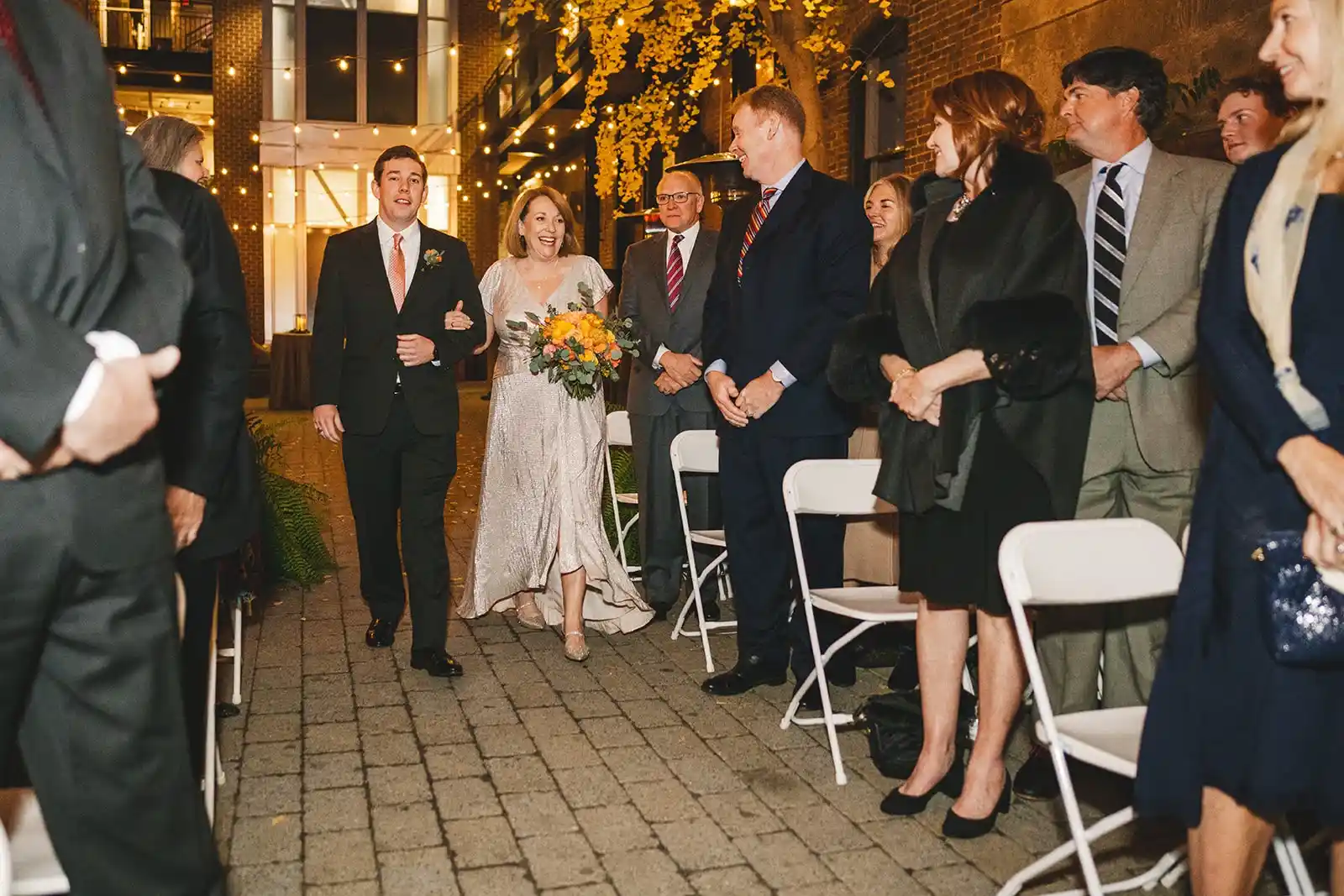 Bride in a silver gown holding a yellow and orange bouquet walks down the aisle with her son during an evening wedding ceremony with string lights.
