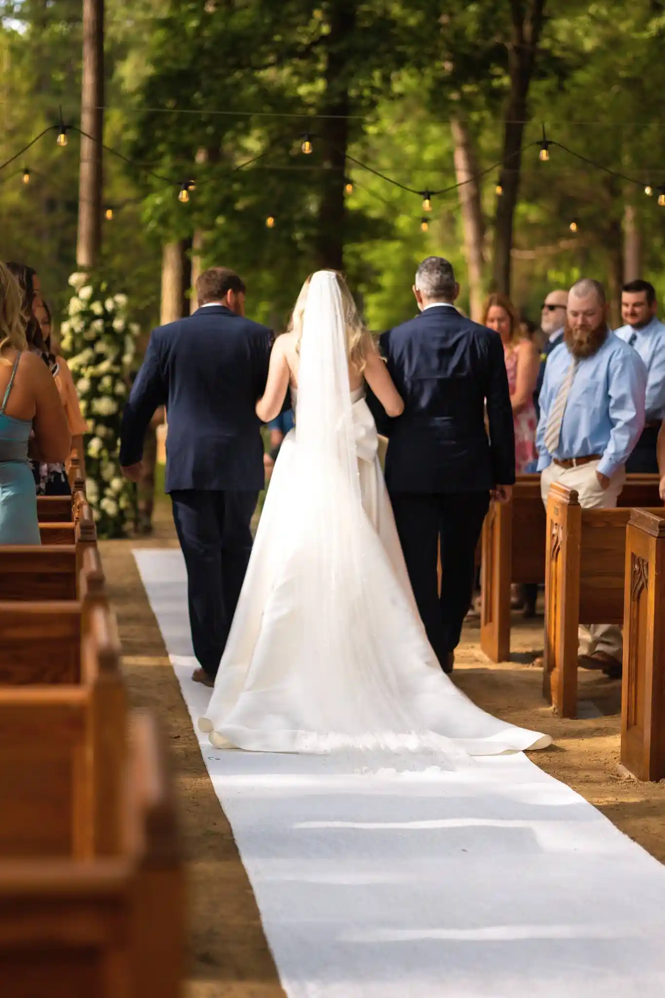 Bride in a white gown and veil walks down an outdoor aisle with two escorts, surrounded by wooden pews and string lights.